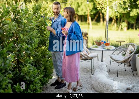 La giovane famiglia passa il tempo estivo, potando cespugli nel cortile della casa di campagna Foto Stock