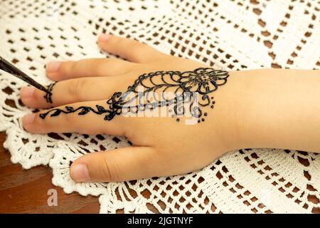 Una ragazza disegna i modelli dell'henné sulla mano di un bambino contro una tovaglia bianca durante una fiera scolastica. Foto Stock