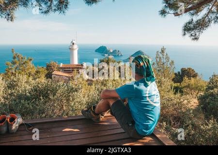 Il viaggiatore siede sullo sfondo di un pittoresco paesaggio marino con piccole isole e un vecchio faro bianco sulla penisola. Un giovane è trekking Foto Stock