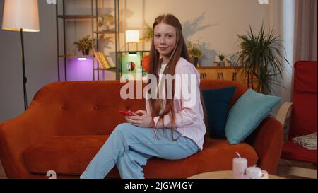 Giovane bambina adolescente seduta sul divano usa il sorriso del telefono cellulare in casa appartamento moderno. Ginger kid texting Condividi i contenuti dei messaggi sulle applicazioni social media per smartphone online, guardando un film rilassante Foto Stock