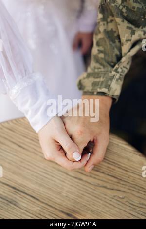 Sposa in abito bianco e sposo in uniforme militare si tengono le mani. Primo piano. Concetto di matrimoni durante la guerra. Guerra in Ucraina. Foto Stock