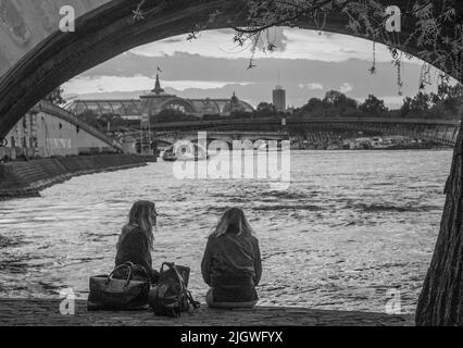 05+-15-2016 Parigi, Francia. 2 giovane donna seduta sulla riva della Senna e tramonto e Grand Palace (Palais) in lontananza. Possono essere turisti(?). Foto Stock