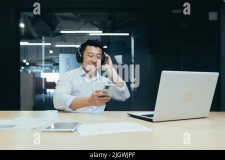 Uomo d'affari in ufficio in pausa di lavoro, ascolto di musica in grandi cuffie nere, uomo asiatico utilizzando l'app musicale sul telefono Foto Stock