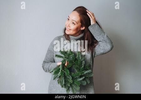 Donna sorridente adulta quarant'anni con capelli ricci brunette in caldo abito a maglia grigio con corona di natale in abete fai da te in mano su sfondo grigio Foto Stock