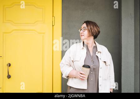 Donna di età media brunetta per adulti più corpo di taglia positivo in abiti casual con tazza di carta di caffè vicino alla porta gialla Foto Stock