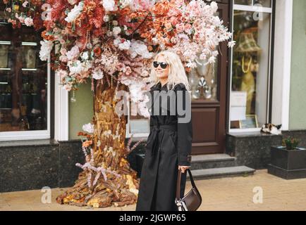 Giovane donna bionda alla moda in un impermeabile nero cammina attraverso i negozi della città Foto Stock