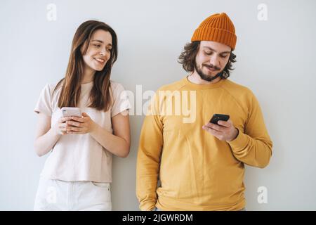 Coppia i giovani hipsters della famiglia che usano i telefoni cellulari sullo sfondo grigio della parete Foto Stock