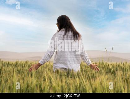 Donna contadina, capelli lunghi casual vestito donna contadina. Donna agronomista in piedi nel campo di grano verde. Orgoglioso della sua fattoria. Toccare le orecchie di grano Foto Stock