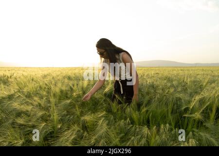Donna contadina, moderna donna contadina dai capelli lunghi. Donna agronomista casual vestita in piedi nel campo di grano verde. Controllando i suoi raccolti. Foto Stock