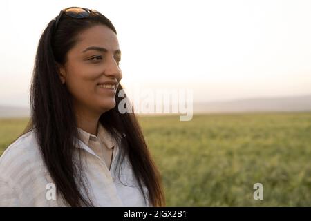 Donna agricoltore, ritratto di vista laterale di giovane donna agricoltore in cerca di spazio copia. Agronomo femminile in piedi e sorridente nel campo di grano verde. Foto Stock