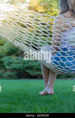 Vista verticale di una giovane donna irriconoscibile seduta in un'amaca con le gambe appesi in estate nel parco del campeggio Foto Stock