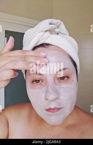 Candid donna applica maschera di argilla, minerali e sali al suo viso in bagno di casa. Cura della pelle di auto durante il covid-19, donna reale, momento di lifestyle candid Foto Stock