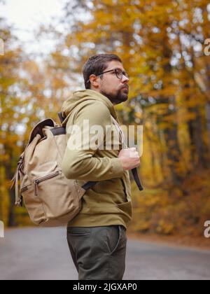 Giovane bel uomo in posa in autunno foresta su strada. Giovane hipster ragazzo con zaino , viaggiatore in piedi in legno, Escursionismo, Foresta, viaggio, attivo sano stile di vita, avventura, concetto di vacanza. Foto Stock