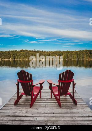 Due sedie rosse adirondack alla fine di un molo che si affaccia su un lago. Foto Stock