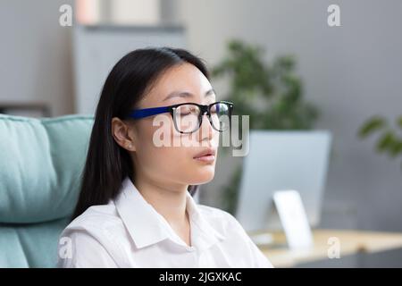Pausa di riposo al lavoro. Giovane bella donna asiatica in occhiali seduti alla scrivania in ufficio con occhi chiusi riposanti, rilassato, meditating. Foto Stock