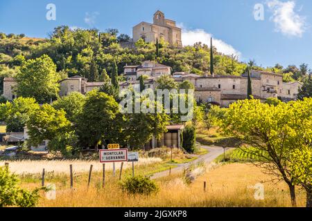 Case in pietra a terrazze si arrampicano sulla collina boscosa di Montclar-sur-Gervanne, nella Drôme, Francia. La chiesa romanica di Saint-Marcel del XII secolo si affaccia sul villaggio. Cipressi fiancheggiano il nucleo storico, un tempo protetto dalla muraglia circolare "vingtain" con due torri rotondi e il portale Porte Bayard. Ai piedi della collina si trova il cartello stradale sulla D 577. Foto Stock