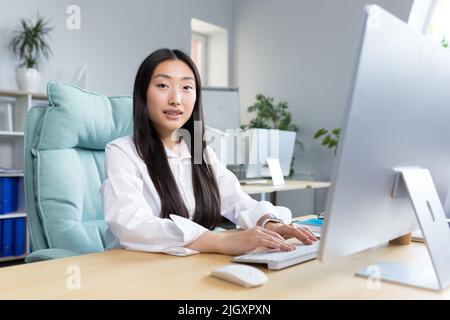 Foto da primo piano. Ritratto di una giovane bella donna asiatica segretario seduto alla scrivania al computer in ufficio in abiti da lavoro, guardando la macchina fotografica, sorridente. Foto Stock