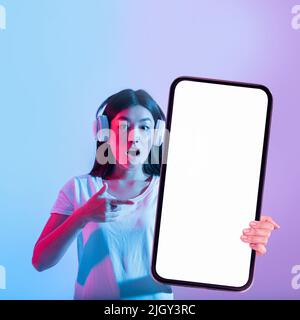 App cool. Scioccata Asian Female in cuffie che puntano a Big smartphone vuoto Foto Stock