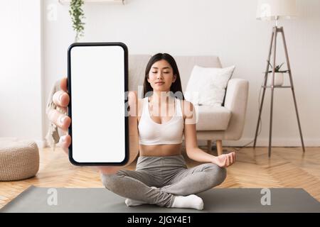App Yoga. Calma giovane donna asiatica meditating e tenendo grande smartphone vuoto Foto Stock