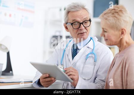 Medico senior con gravi rughe in camice da laboratorio seduto al tavolo e utilizzando un tablet digitale con i record dei pazienti online, dando consigli a matu Foto Stock