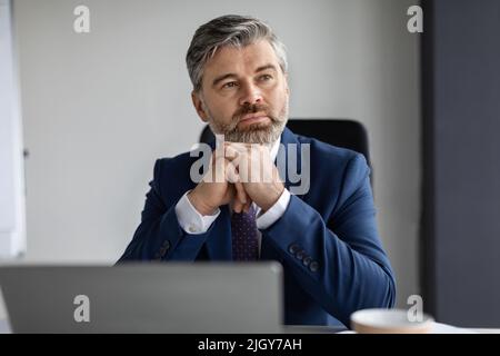 Pensieroso imprenditore di mezza età seduto alla scrivania in ufficio e guardando via Foto Stock