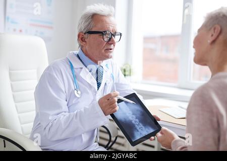 Fidato medico di sesso maschile senior in camice da laboratorio utilizzando tablet digitale mentre si parla al paziente di disturbi del midollo spinale Foto Stock