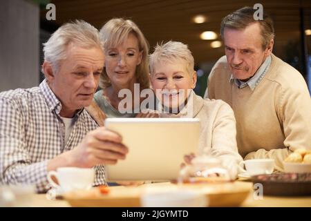 Gruppo di curiosi anziani ex compagni di classe in abbigliamento casual seduti a tavola e guardando vecchie foto sul tablet Foto Stock