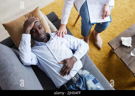 Vista ad angolo alto del terapeuta femminile con appunti sulle ginocchia che toccano la spalla del paziente nero mentre lo confortano durante la sessione di terapia Foto Stock