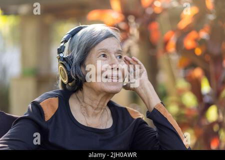 Donna asiatica anziana che ascolta musica con cuffie nel cortile. Foto Stock