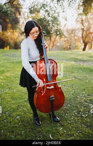 Giovane donna bruna in piedi con occhiali che suonano violoncello al tramonto nel parco, su un'erba verde. Foto Stock