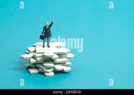Fotografia di personaggi giocattolo in miniatura. Un uomo d'affari che si alza sopra il mucchio di pezzi di puzzle jigsaw mentre alza la mano. Isolato su sfondo blu. Imag Foto Stock