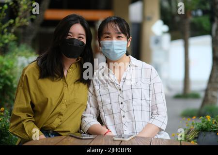 Due giovani donne asiatiche in maschera di viso seduti al caffè sul marciapiede, guardando la macchina fotografica Foto Stock