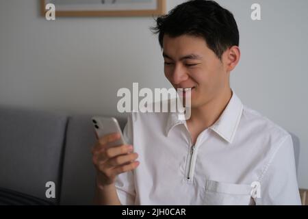 Bel giovane asiatico che guarda lo smartphone con sorridente a casa Foto Stock