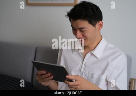 Bel giovane asiatico che guarda il tablet con sorridente a casa Foto Stock