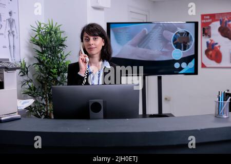 Ritratto di un sorridente receptionist ospedaliero che lavora alla reception telefonicamente parlando con il paziente che fa un appuntamento. Donna dall'aspetto professionale che risponde alla chiamata telefonica in clinica privata. Foto Stock