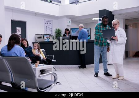 Medico senior che tiene un laptop che presenta i risultati di laboratorio a un paziente afroamericano in clinica privata. Vecchio medico che parla con l'uomo che controlla l'appuntamento sul computer portatile. Foto Stock