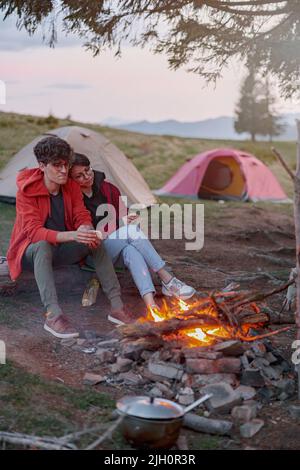 Coppia romantica caucasica seduta al fuoco e riscaldamento in abbracci vicino tende. Riposa in natura. Foto Stock