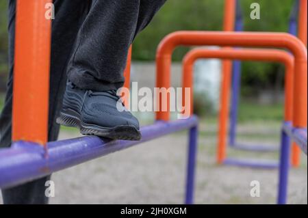 La parte inferiore di un uomo maturo impegnato in ginnastica rigenerativa sul terreno sportivo. Le gambe dell'uomo sono in piedi sulle barre blu e arancione. Reg Foto Stock