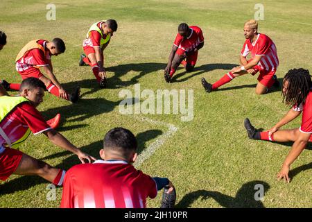 Vista ad alto angolo di atleti maschi multirazziali che allungano le gambe sul campo erboso in estate parco giochi Foto Stock