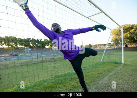 Portiere di sesso maschile afro-americano con le braccia sollevate che saltano e catturano la palla di calcio durante la partita Foto Stock