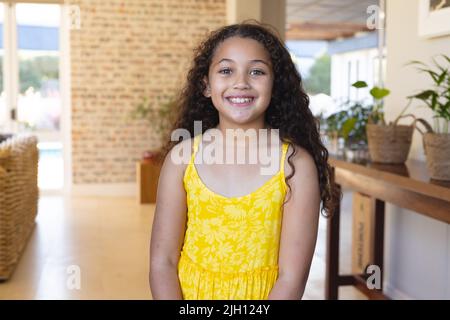 Ritratto di sorridente ragazza simpatica biraciale con capelli ricci lunghi vestito giallo in piedi a casa Foto Stock