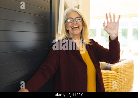Ritratto di allegra anziana caucasica ridendo e ondeggiando la mano mentre si dà il benvenuto all'ingresso Foto Stock