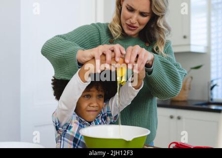 Madre multirazziale che fa il viso mentre aiuta il figlio con capelli afro in rottura uovo in ciotola in cucina Foto Stock