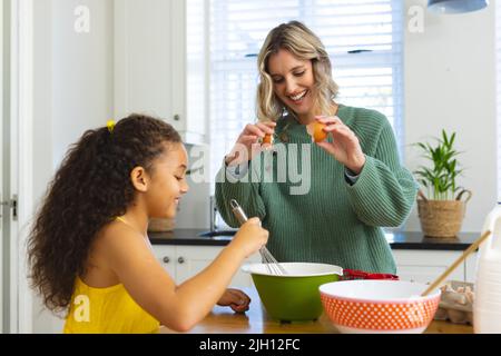 Multirazziale giovane madre che rompe l'uovo mentre la figlia che sbatte la pastella in una ciotola in cucina Foto Stock