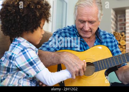 L'anziano caucasico insegna a suonare la chitarra al nipote biraciale mentre si siede a casa Foto Stock