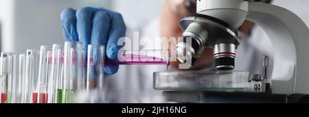 Scienziata femminile che versa il liquido del campione rosa su un contenitore di vetro al microscopio in laboratorio Foto Stock