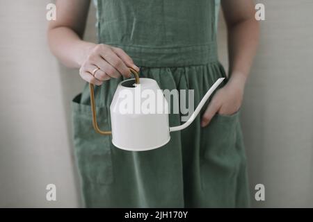 Ragazza in grembiule tiene un annaffiatoio. Annaffiatura e cura di piante domestiche. Giardinaggio domestico. Coltivazione di fiori di hobby Foto Stock