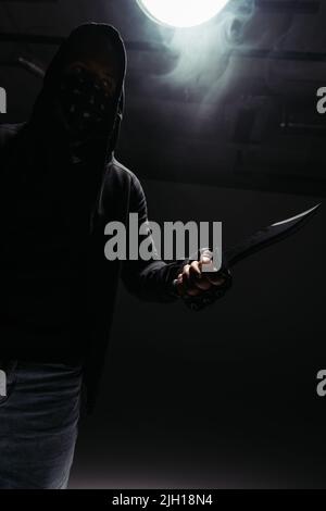 Silhouette di hooligan afroamericano con faccia oscura che tiene coltello su sfondo nero con fumo Foto Stock