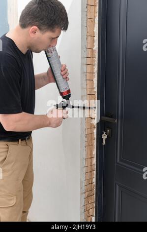 un uomo usa una pistola a spruzzo con la mano e riempie lo spazio con schiuma da costruzione tra una nuova porta di legno e un muro. Foto Stock