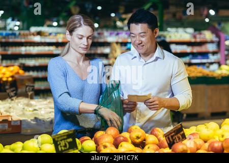 felice misto razza asiatica coppia uomo e donna famiglia scelta frutta o verdura nel supermercato negozio di alimentari. Shopping quotidiano congiunto. Acquirenti cliente seleziona prodotto scegliere una mela in borsa ecologica Foto Stock